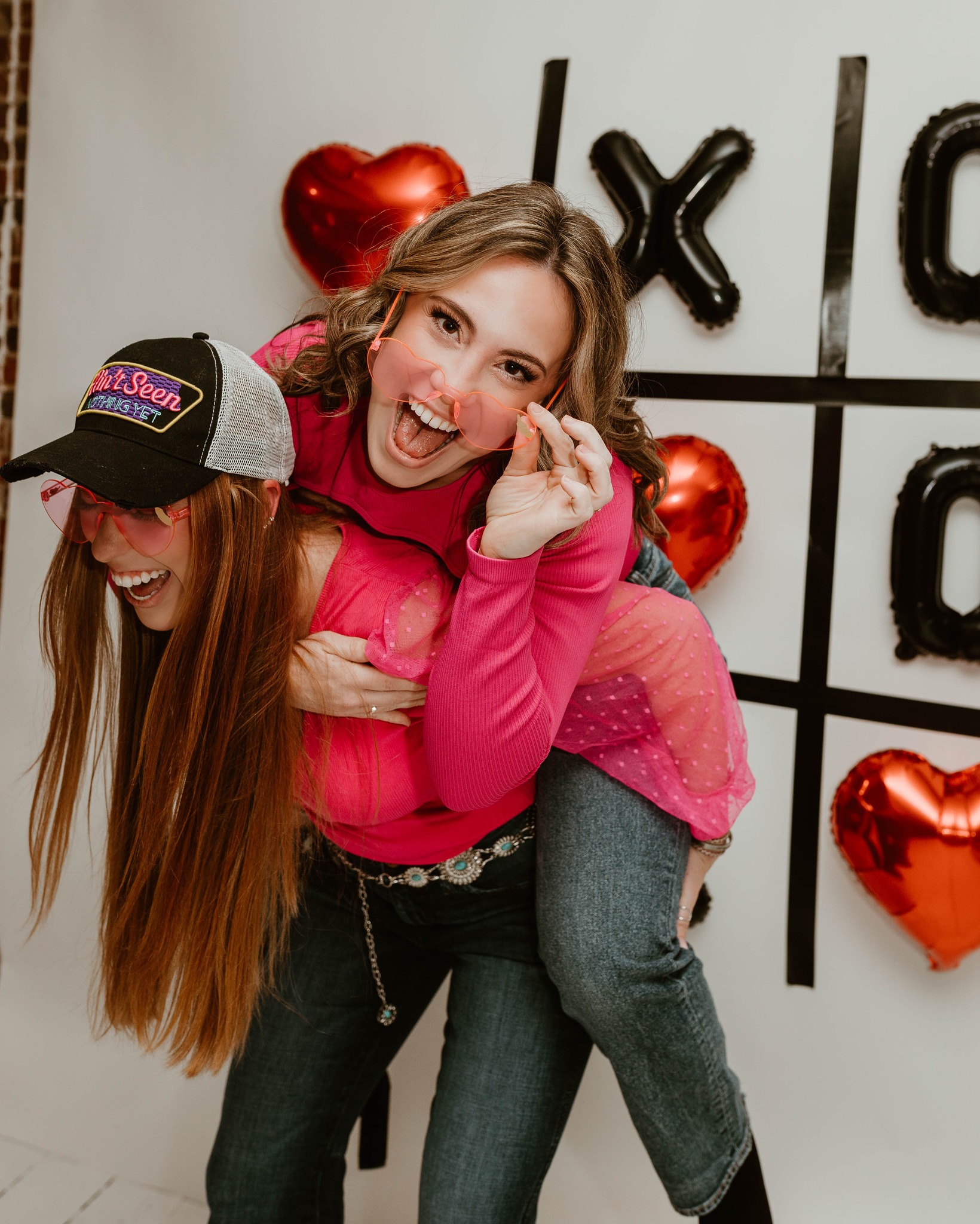 two female friends in front of tic tac toe backdrop. One friend is giving the other a piggy back ride. They are smiling. and wearing heart shaped glasss